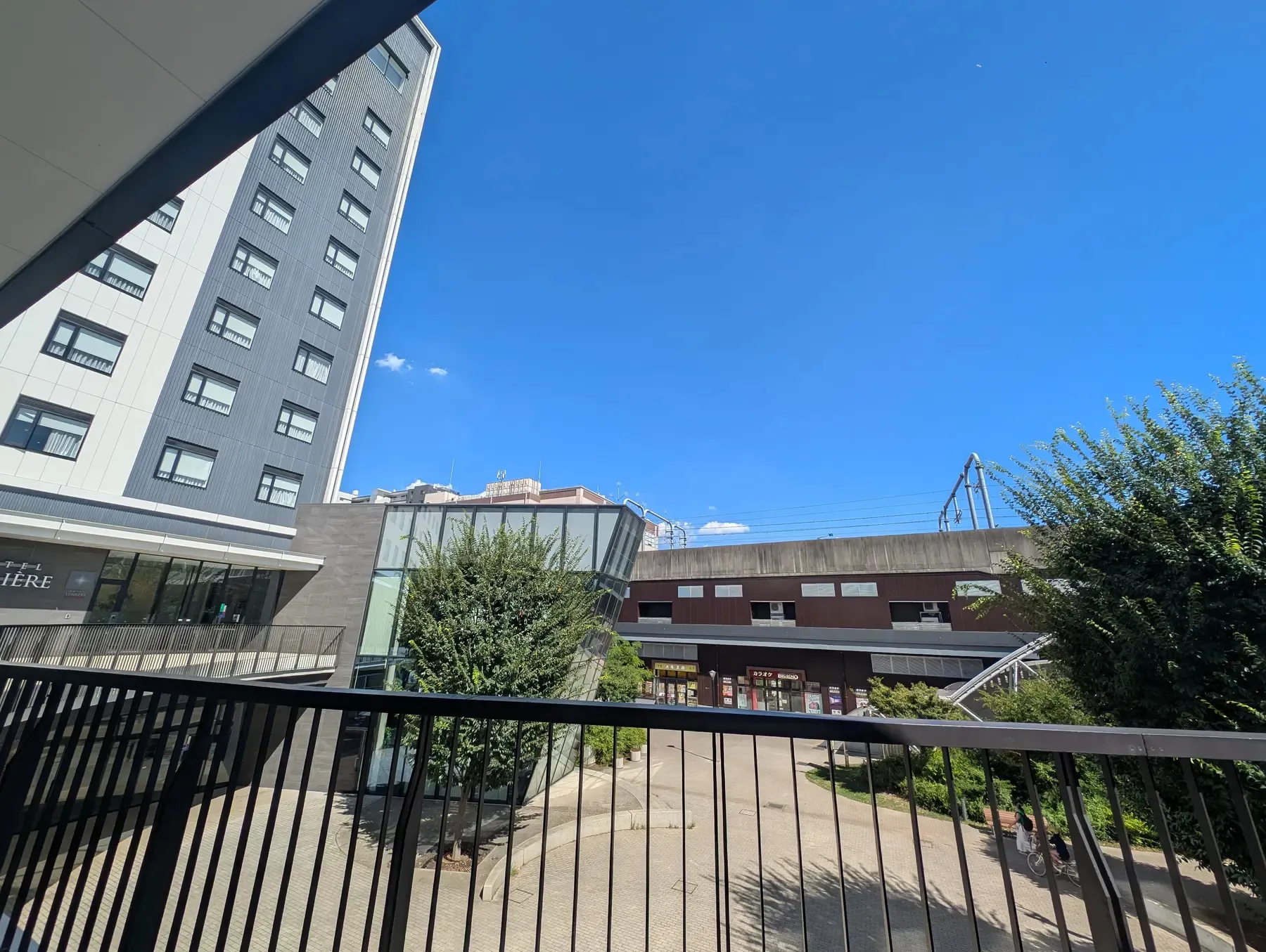 流山おおたかの森駅北出口川の建物群とフェンス越しの緑地。晴れた空の下で街の景観が見える。